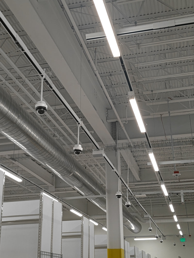Industrial interior scene with track busway runs suspended below the ceiling, supporting linear LED lights and security cameras, alongside exposed ductwork and warehouse shelving.