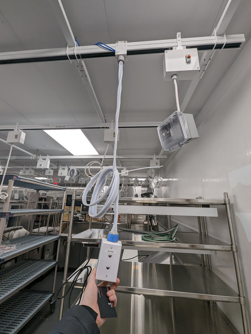 Commercial workspace showing a track busway along the ceiling with several plug‑in power drops and outlet boxes; a person holds a coiled cord beneath stainless‑steel racks.